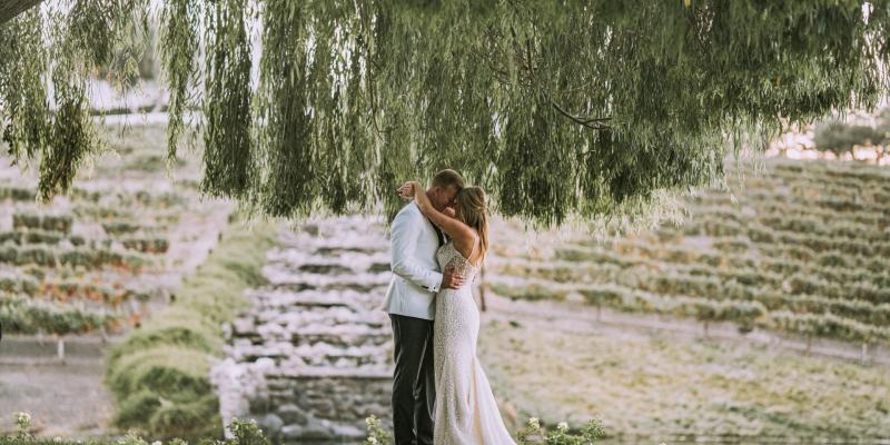 Couple embracing beneath willow trees at a vineyard, showcasing a seamless indoor-outdoor setting for private event venues.